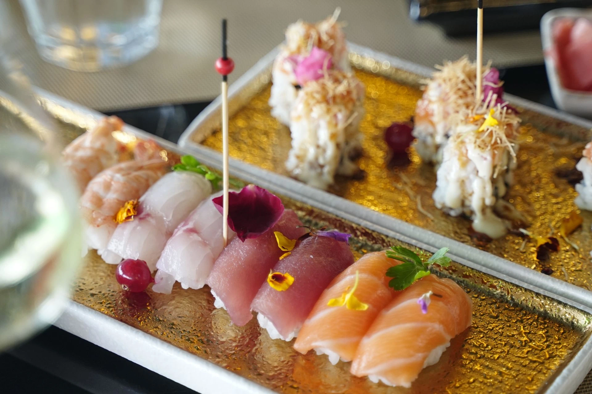 Su un vassoio dorato sono presenti dei sushi giapponesi raffinati, tra cui diversi tipi di sushi con 刺身 (sashimi) di salmone, tonno, gambero e altri, decorati con petali viola, piccoli fiori gialli e bacche rosse, con spiedini di legno inserti. Sullo sfondo c'è anche un bicchiere d'acqua e un altro piatto di sushi.