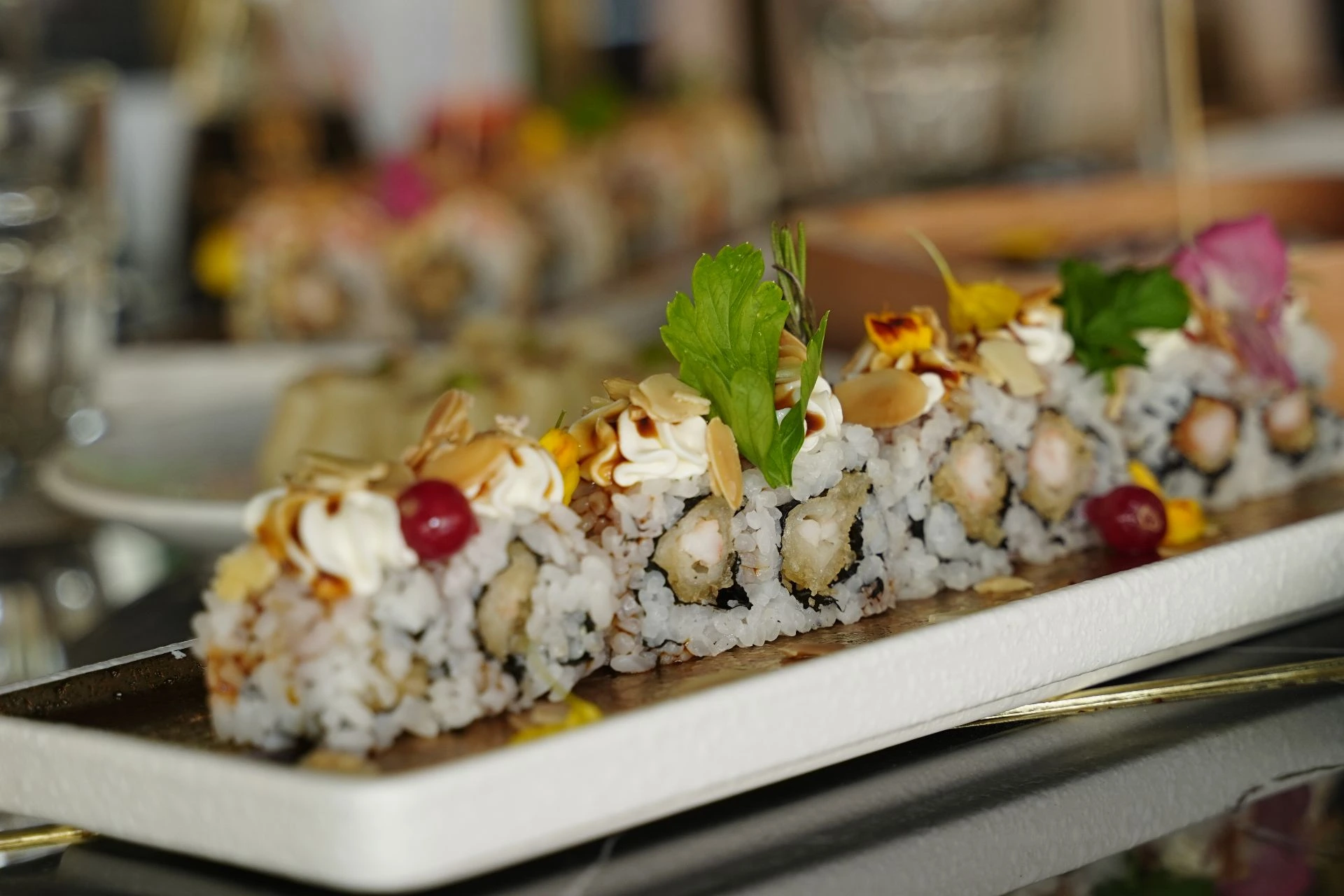 Su un vassoio lungo bianco sono presenti dei rotoli di sushi raffinati, con decori come panna montata, fettine di mandorle, bacche rosse e altri, adornati anche con foglie verdi, ricchi di ingredienti e presentati in modo esteticamente gradevole.